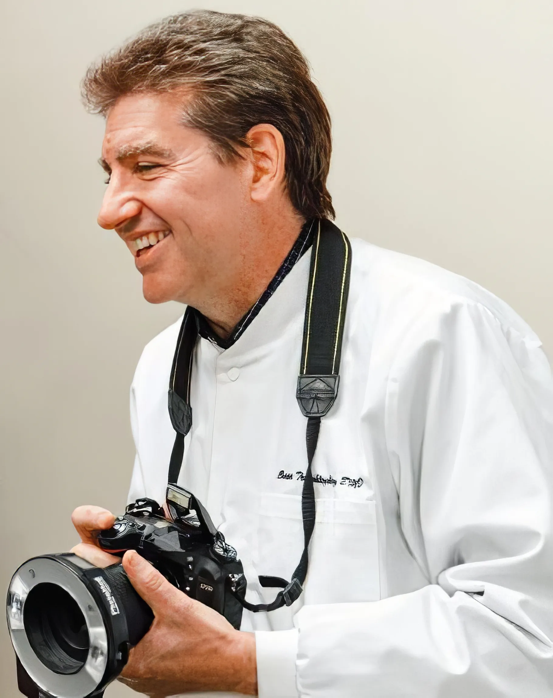 Smiling man in a white chef coat holding a professional camera with a mounted ring light.