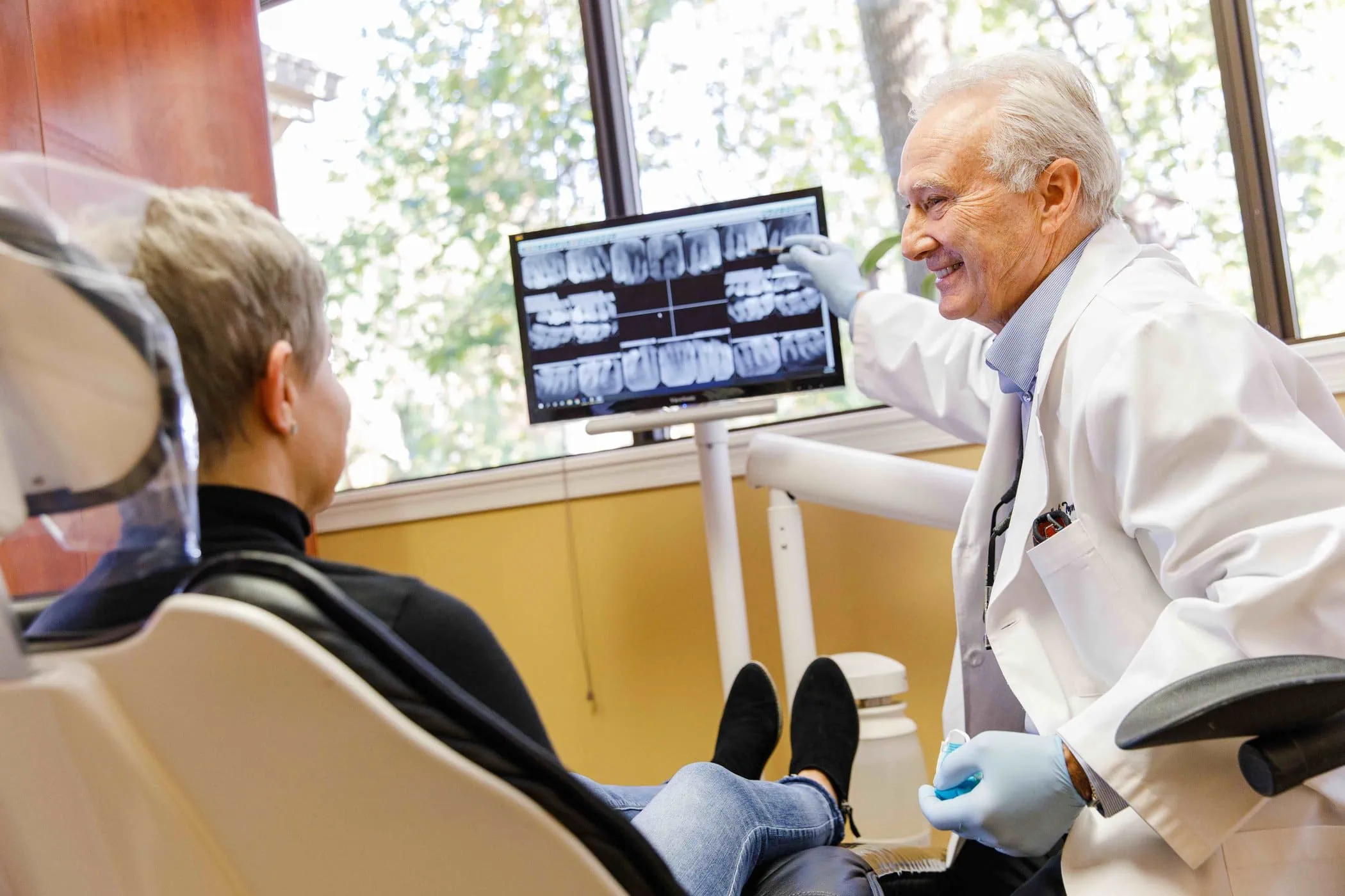 Dentist in white coat explaining dental X-rays on a monitor to a patient seated in a dental chair.