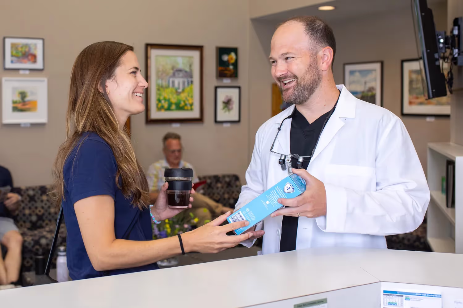 Smiling male dentist in white coat handing a dental product box to a female patient holding a coffee cup at clinic reception.