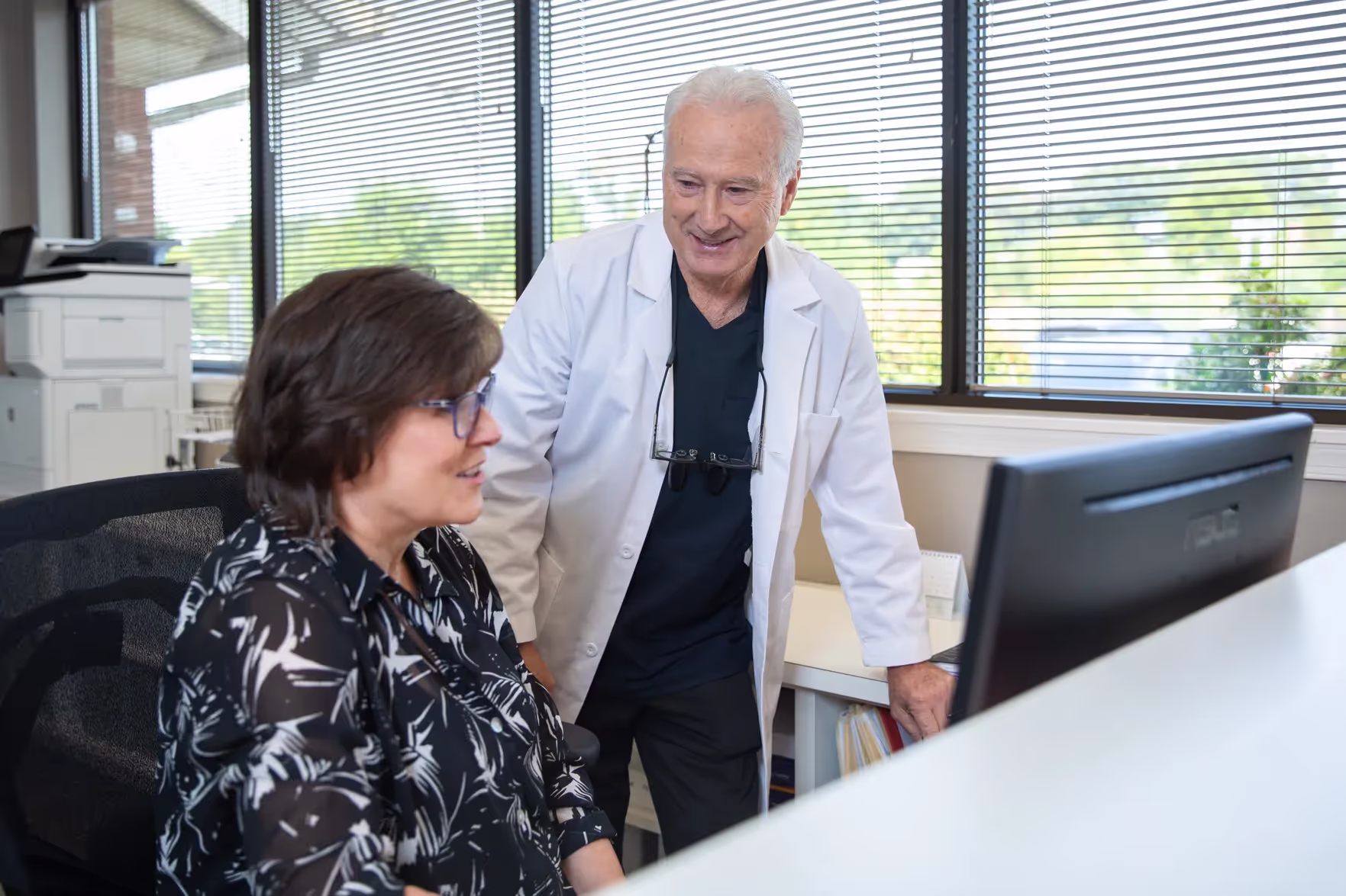 Smiling doctor in a white coat standing beside a seated woman looking at a computer screen in an office with large windows.