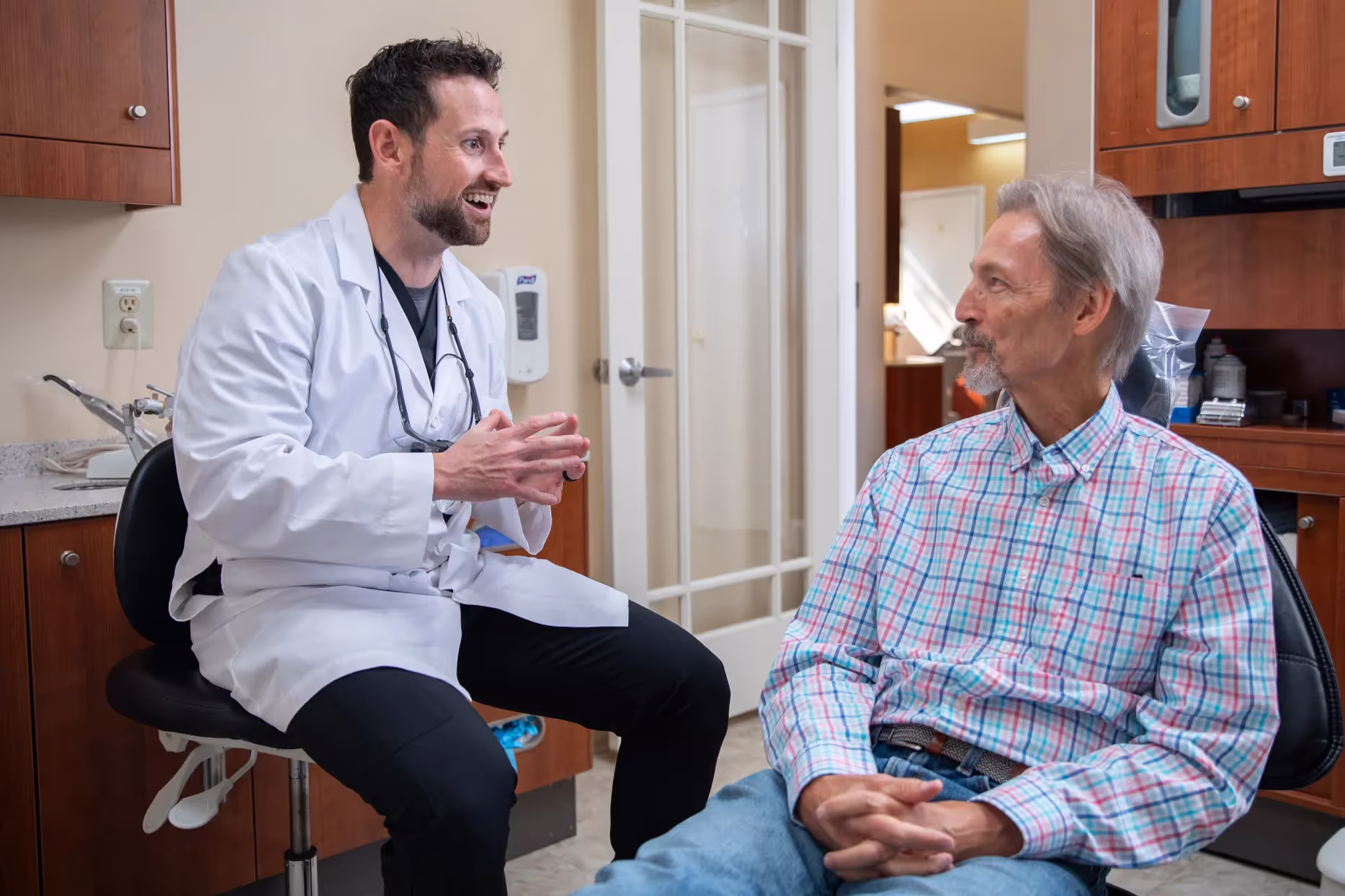Male doctor in white coat talks with male patient seated in medical office.