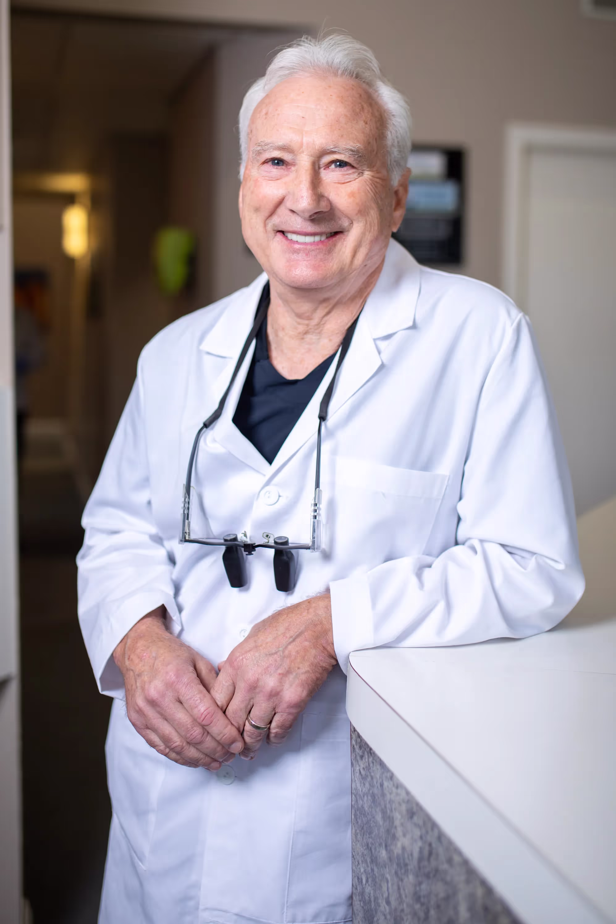 Smiling elderly male doctor in white coat leaning on a counter with medical loupes around his neck.